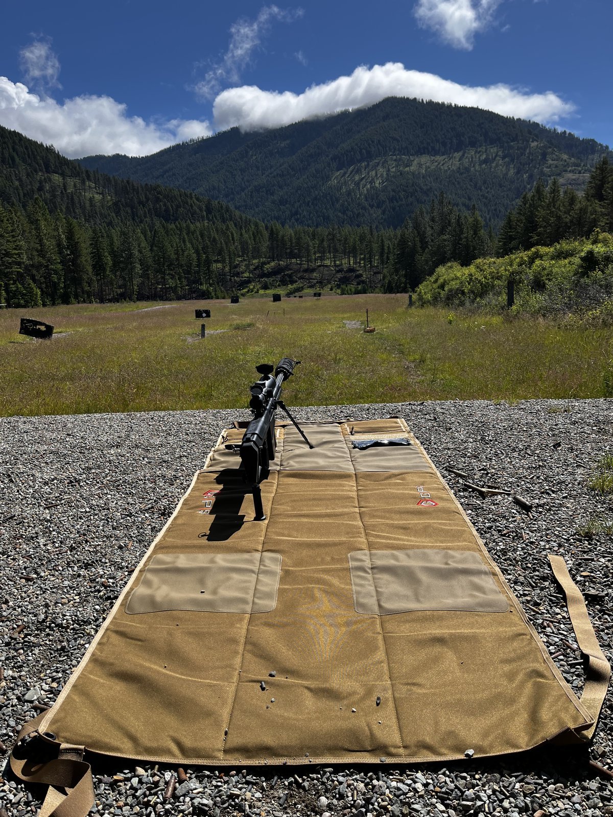 Precision rifle and shooting mat on a gravel range, looking out across a mountain meadow.